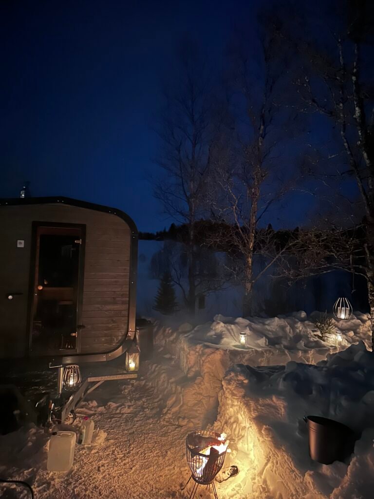Fjellbadstue i vintermørket med bål og snødekt landskap, stemningsfull kveld med varm badstue og flammer som lyser opp snøen.
