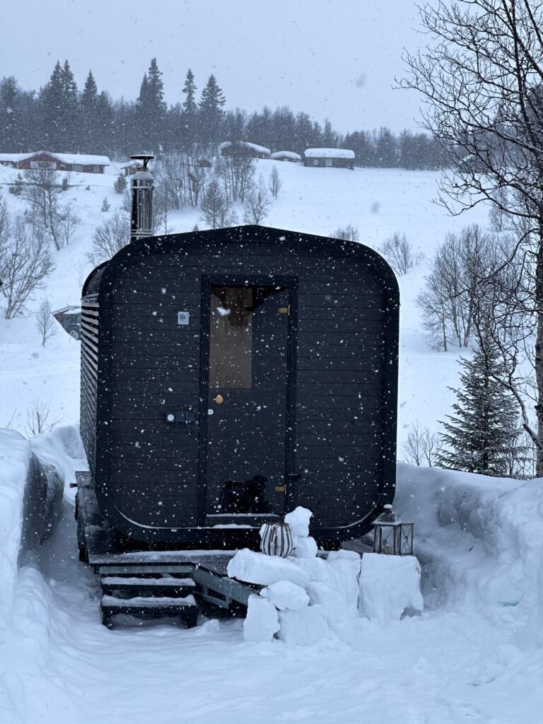 Fjellbadstue omgitt av snø og fjell, med vedfyrt pipe og vinterstemning i fjellandskapet.